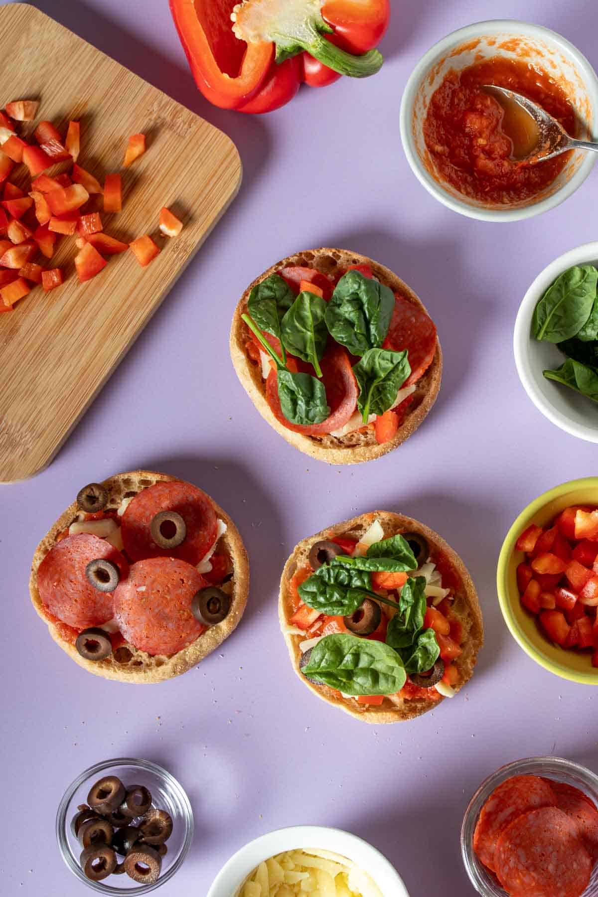 Spinach put on English muffin pizzas topped with pepperoni and veggies and other ingredients on a purple surface next to them.