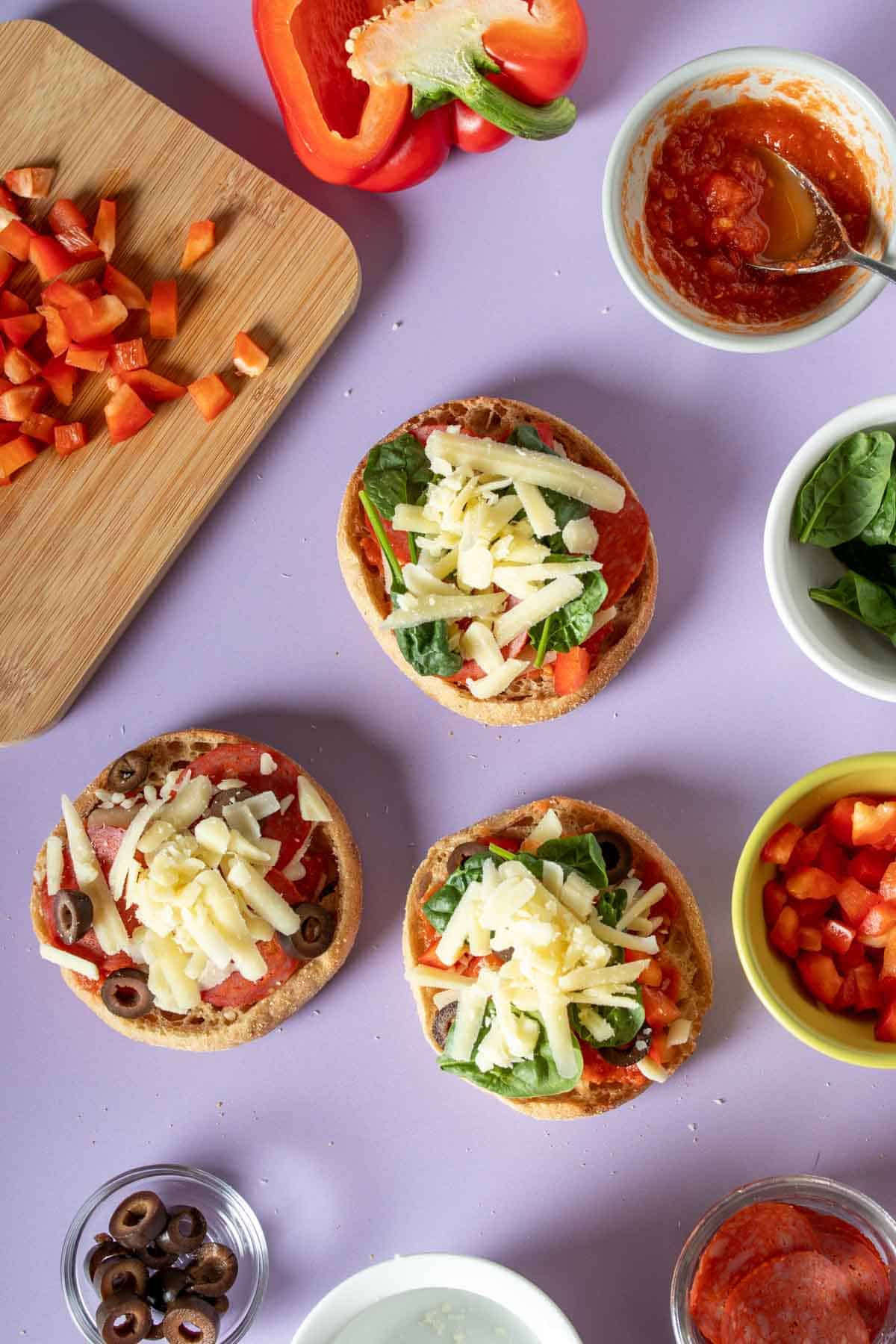 Cheese on the top of English muffins being made into pizzas with veggies and pepperoni and other ingredients around them.