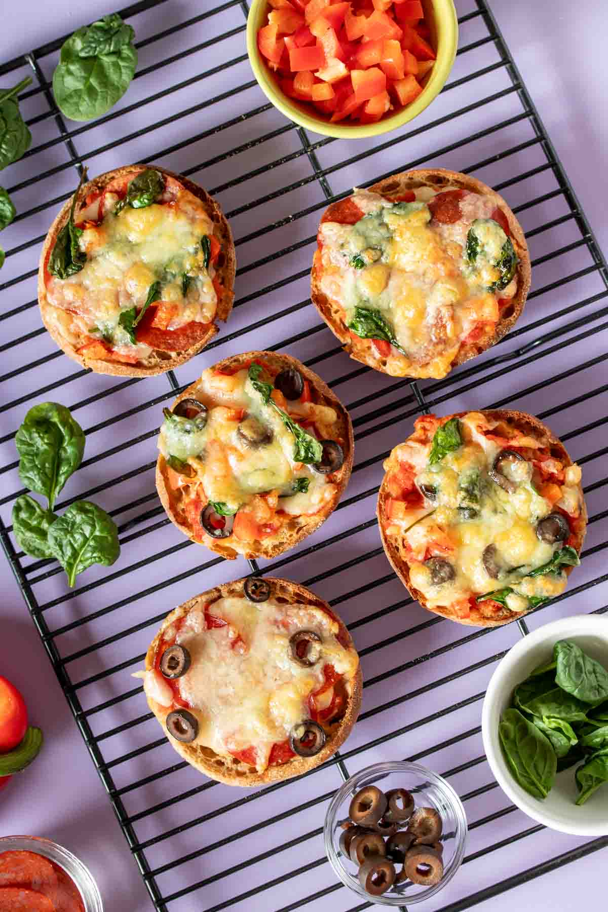Baked English muffin pizzas on a black cooling rack with ingredients in bowls next to them on a purple surface.
