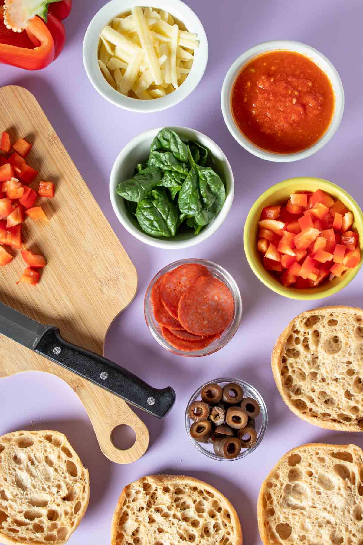 Top view of ingredients and toppings needed to make English muffin pizzas all sitting on a purple surface.