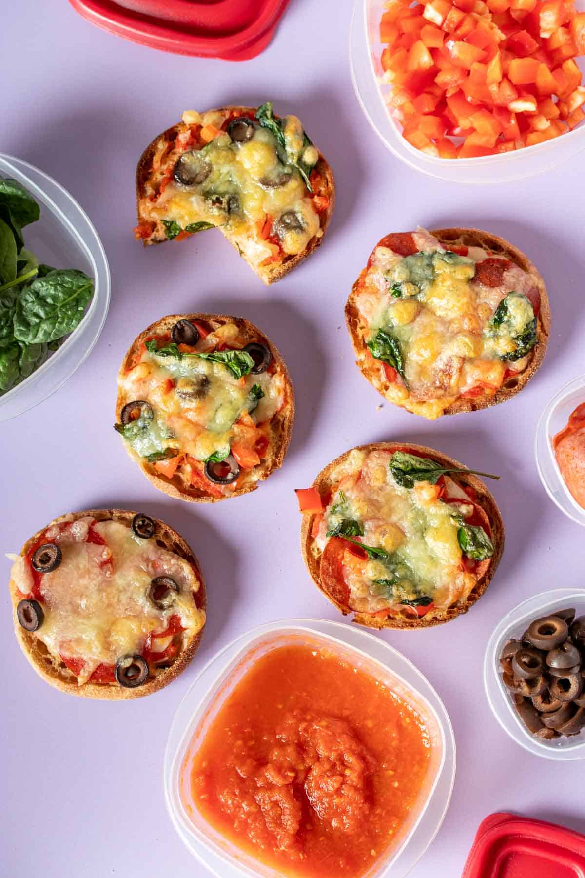 A purple surface with English muffin pizzas and a bite out of one of them and ingredients around them.