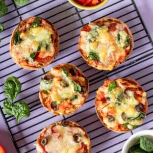 Five English muffin pizzas baked and on a black cooling rack sitting on a purple surface.