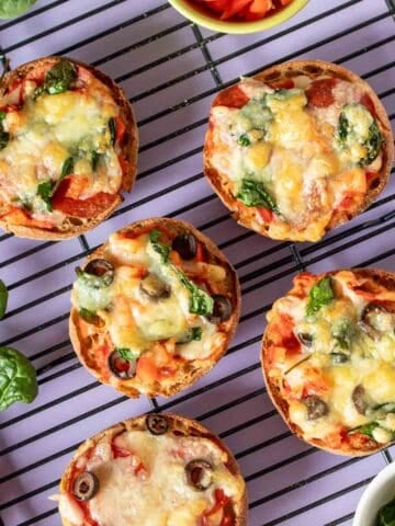 Five English muffin pizzas baked and on a black cooling rack sitting on a purple surface.