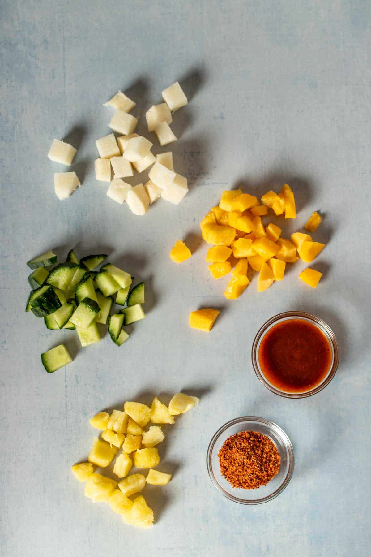 Top view of piles of chopped cucumber, jicama, mango, pineapple and small glass bowls of a red sauce and chile lime seasoning.