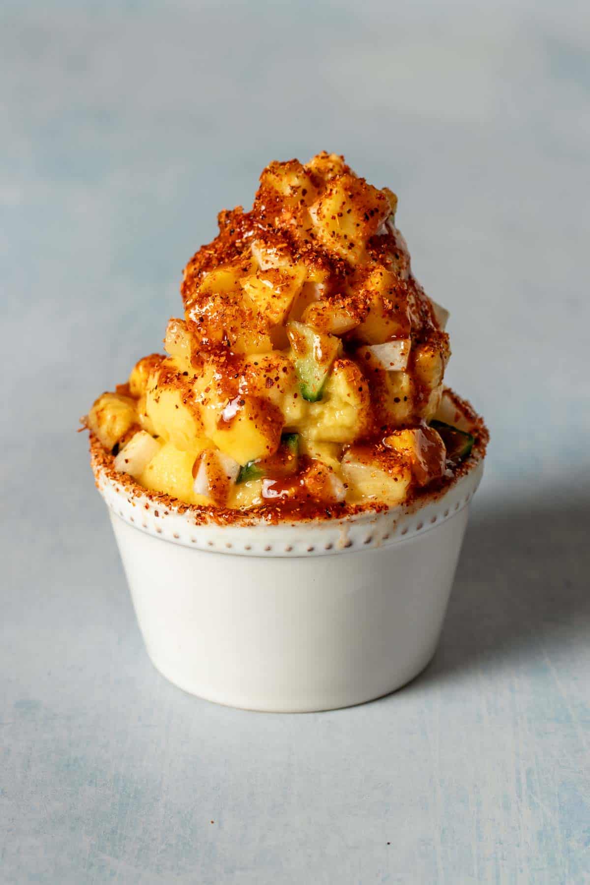 A white bowl with dole whip covered in chopped pieces of fruit and a red sauce with chile lime seasoning.