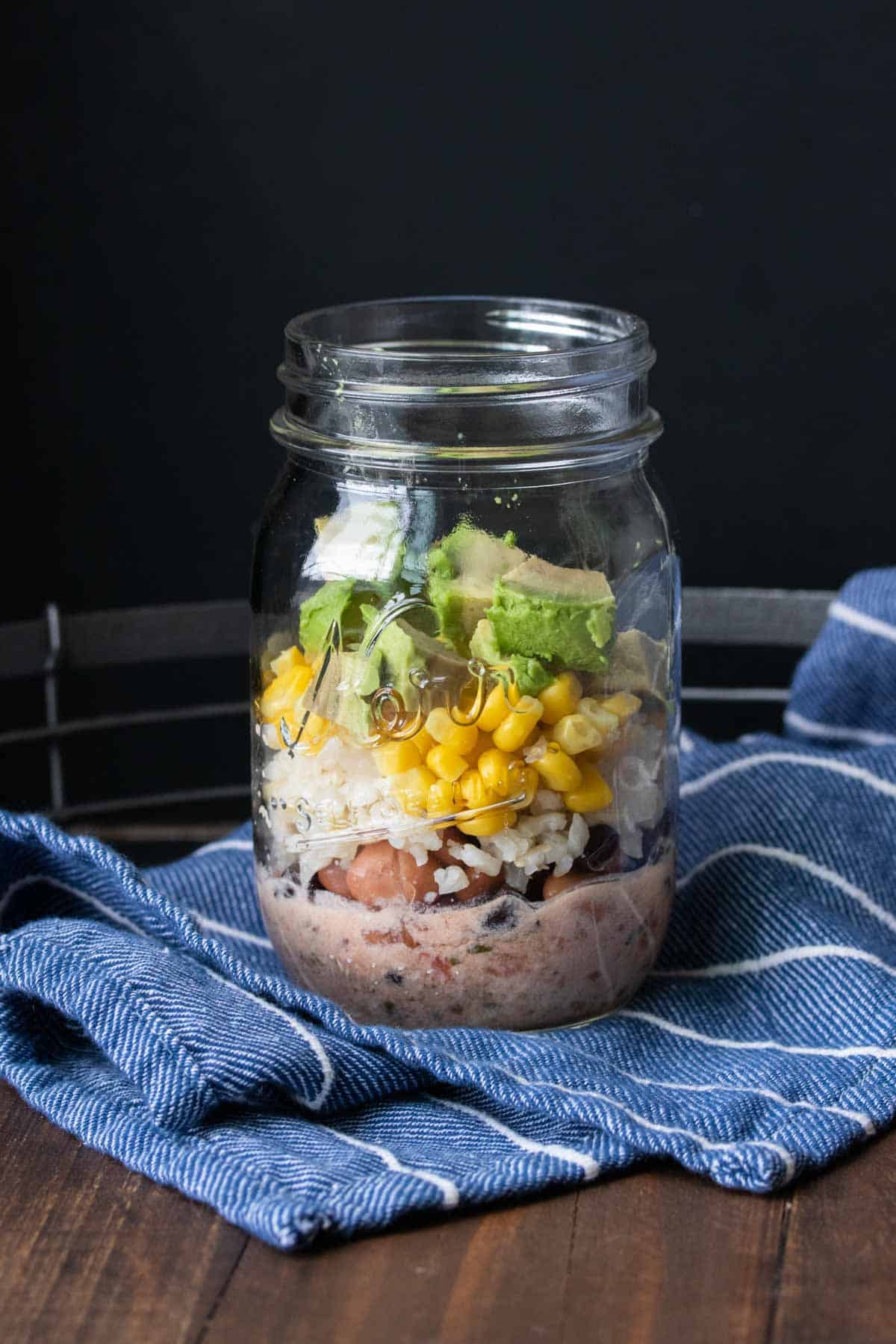 A glass jar on a blue towel filled with beans, rice, corn and avocado.