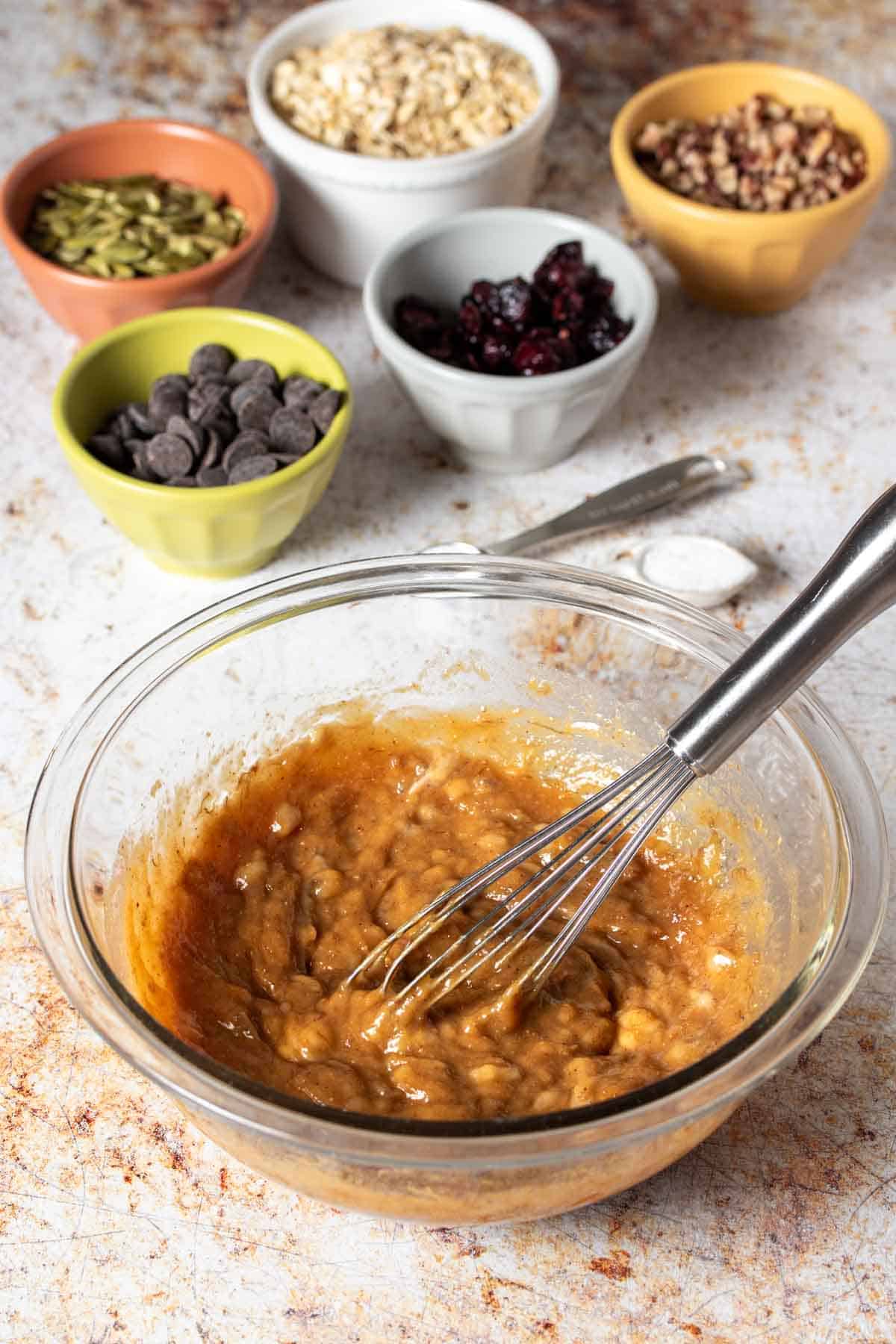 A whisk mixing dough in a glass bowl with ingredients that go in it behind the bowl.