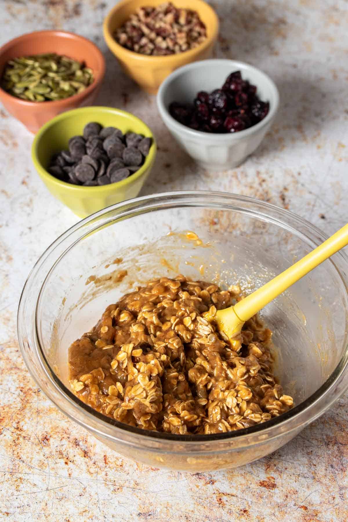 A yellow spatula mixing a dough with oats in a glass bowl in front of toppings.