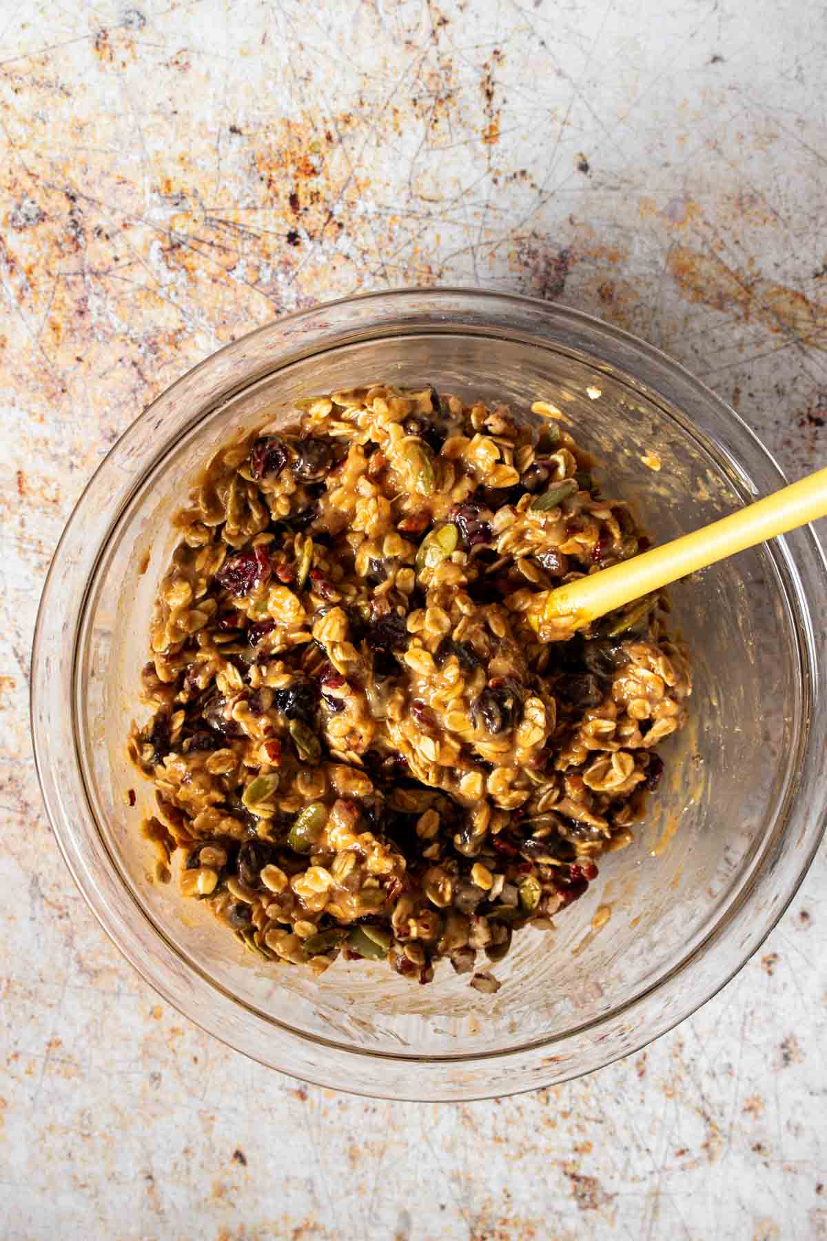 A glass bowl with dough that has oats, nuts, chocolate chips and dried fruit in it.