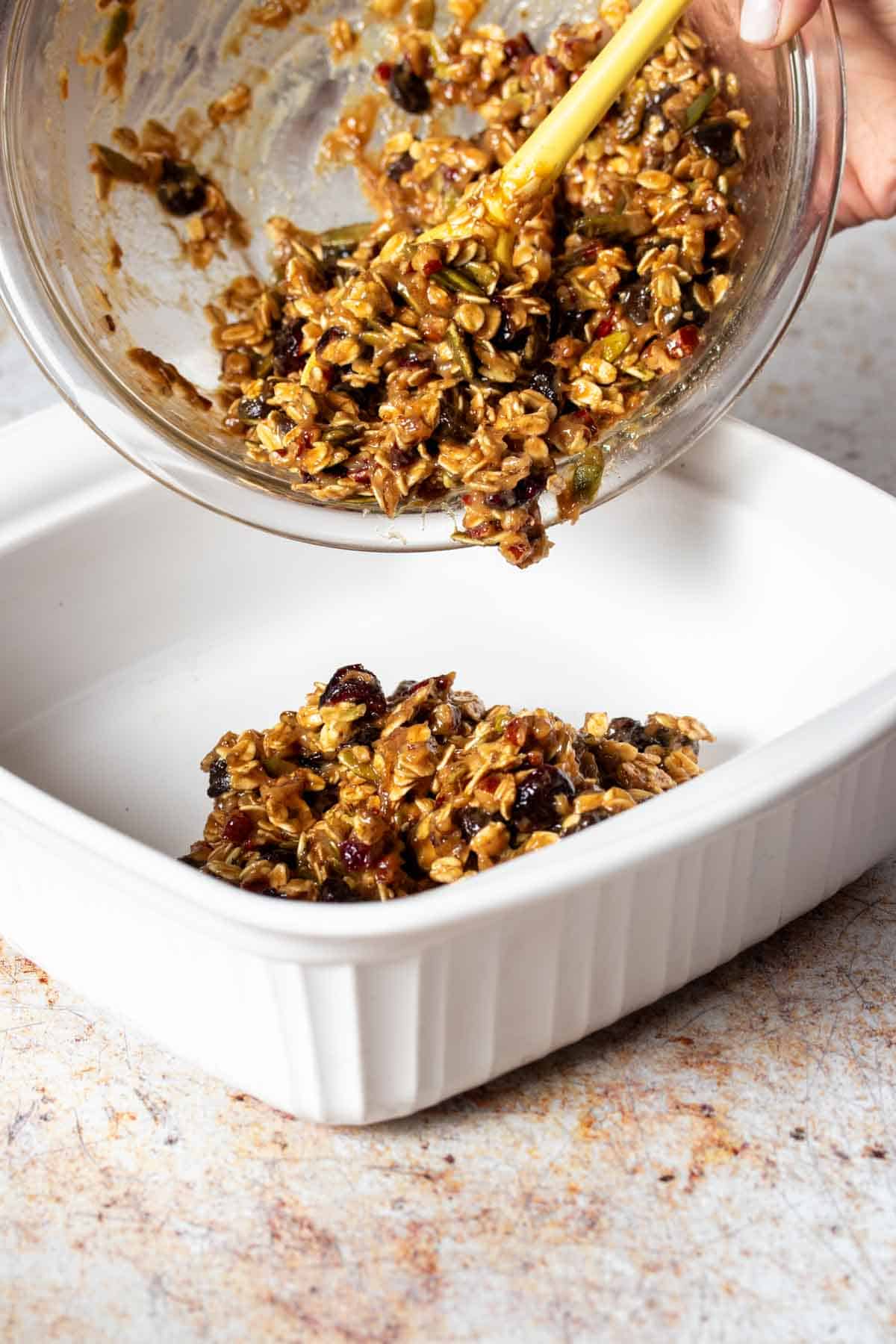 A glass ball with a cookie dough with oats pouring the dough into a white baking dish.