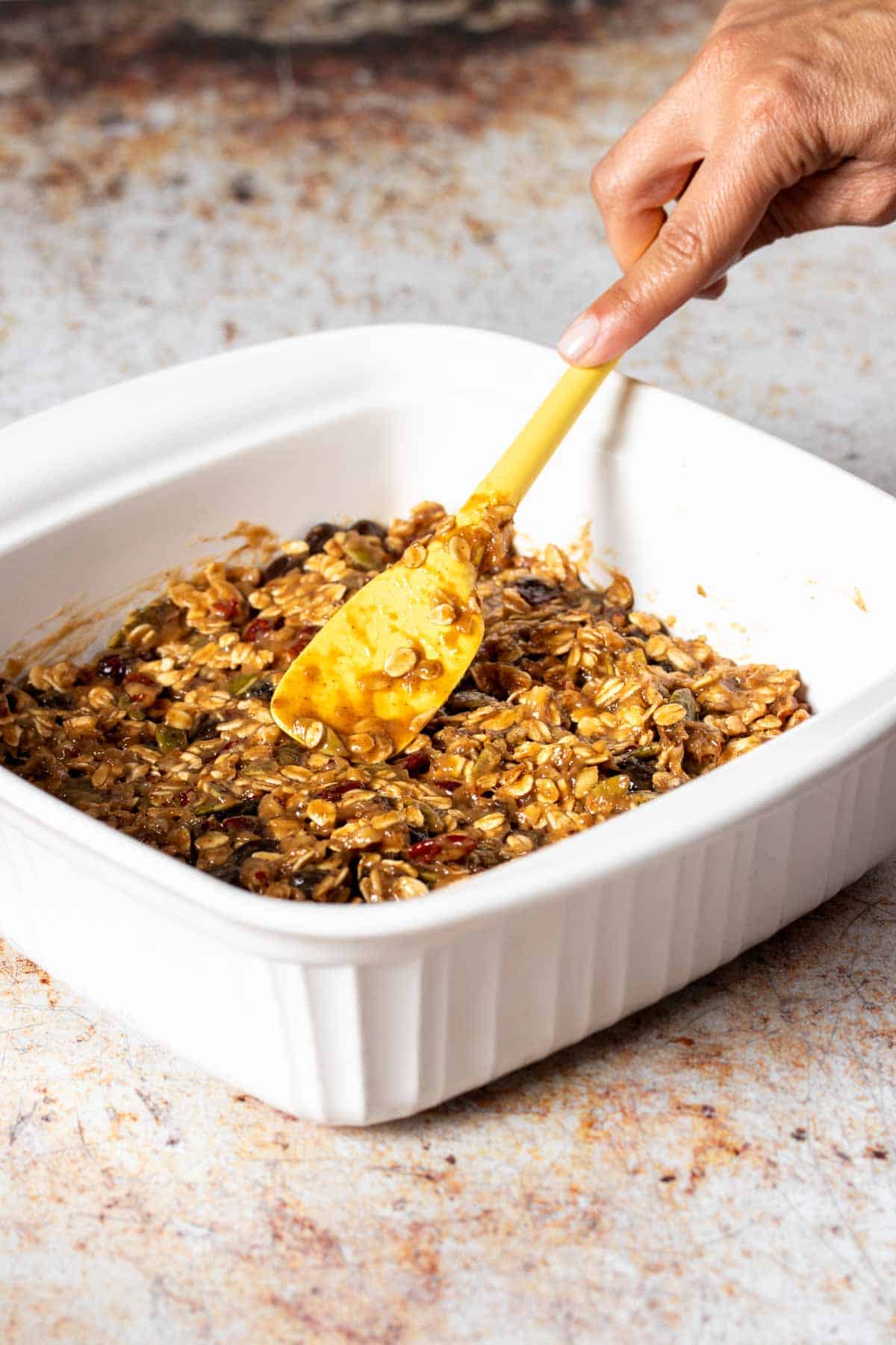 A hand using a yellow spatula to smooth out raw dough with oats in it into a white baking dish.