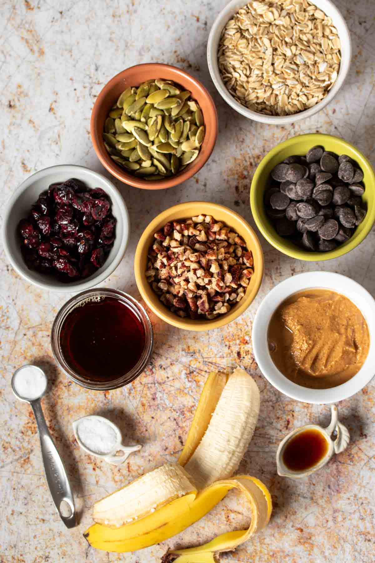 Top view of colored bowls filled with ingredients needed to make oat bars with nuts, dried fruit and chocolate chips with a banana next to them.