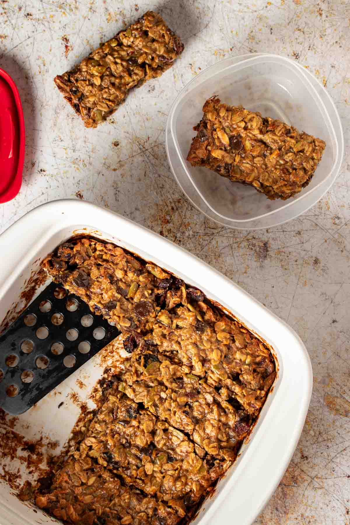 A black spatula putting baked breakfast bars into a plastic container with a red lid.
