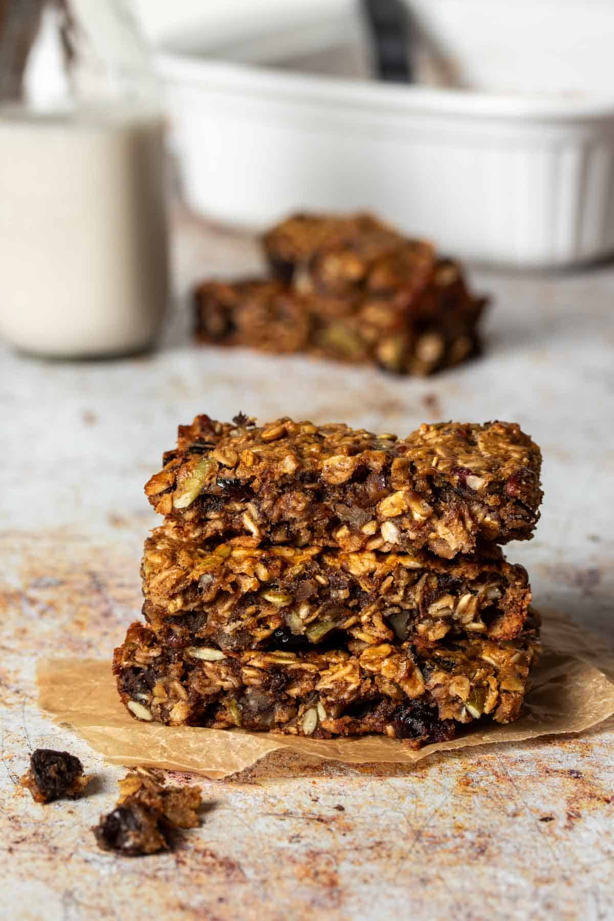 A stack of breakfast bars with oats, nuts and dried fruit sitting in front of more bars, a white pan and a jar of milk.