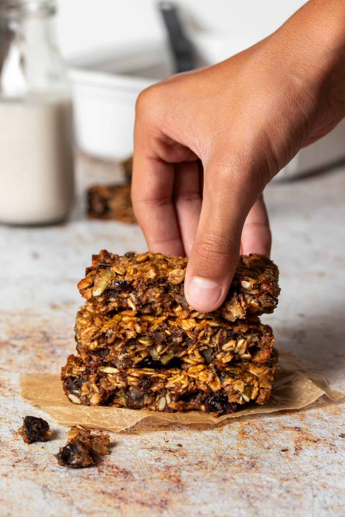 A hand grabbing the top of a stack of three breakfast bars that are on a piece of parchment paper.