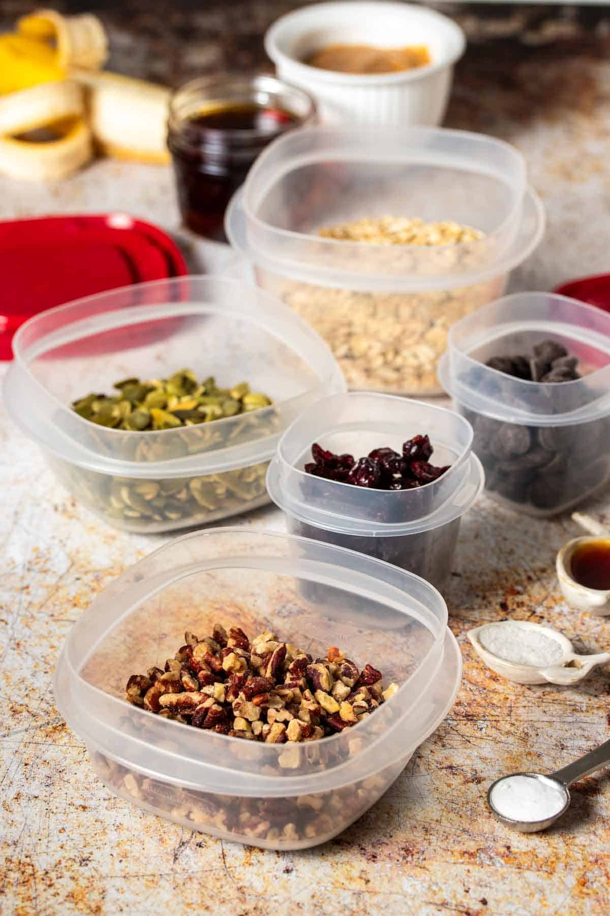 Plastic containers with ingredients to make breakfast bars sitting on a tan speckled surface with red lids next to them.