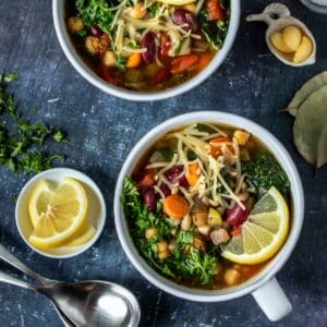 Two white soup bowls filled with vegetable bean soup and sitting on a dark grey surface next to spoons, parsley, garlic and lemons.