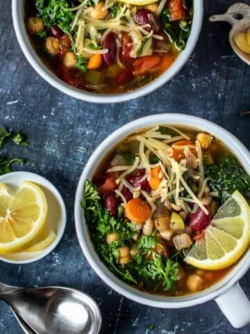 Two white soup bowls filled with vegetable bean soup and sitting on a dark grey surface next to spoons, parsley, garlic and lemons.