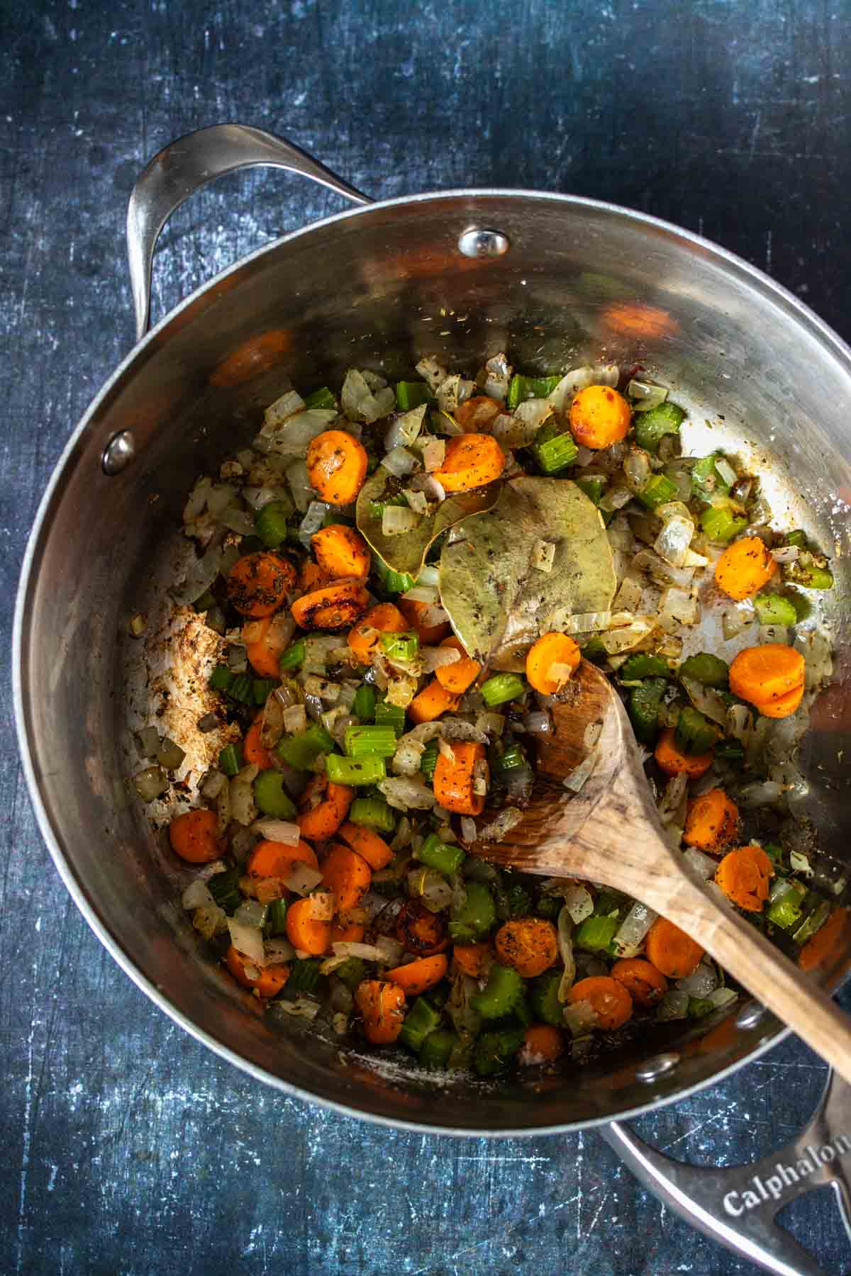 A wooden spoon mixing veggies and spices in a silver soup pot.