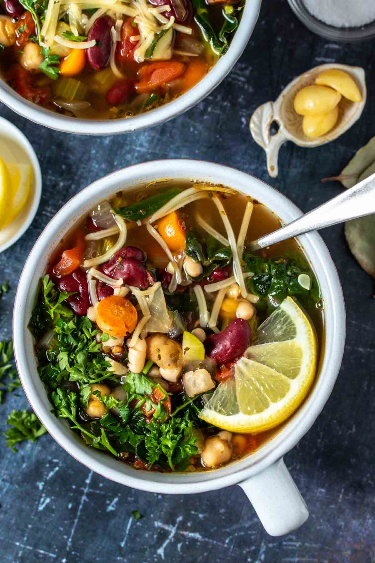 A spoon in a white soup bowl of vegetable bean soup with parmesan and parsley on top with a lemon slice.