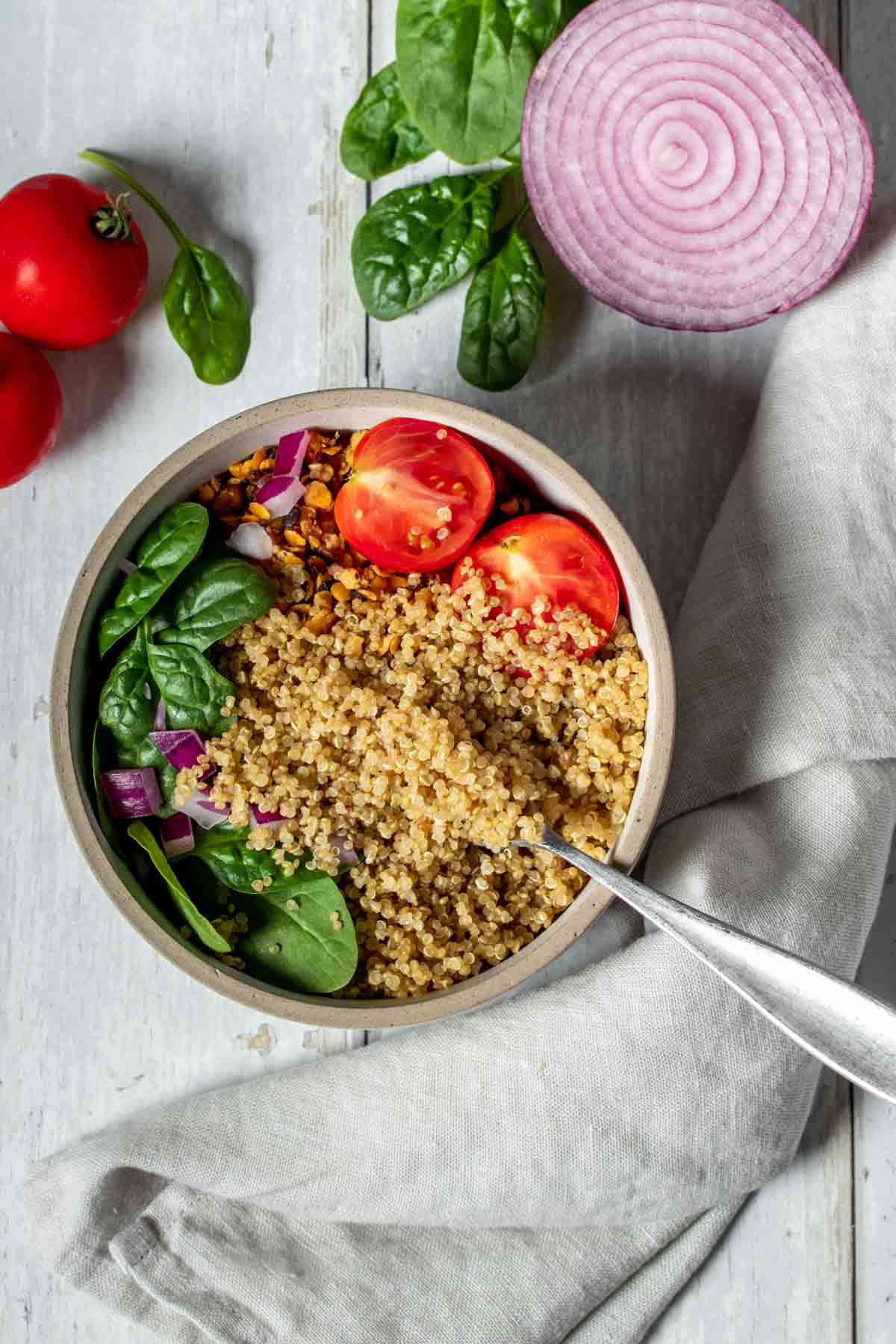 Top view of a white wooden surface with a bowl filled with quinoa, spinach and tomatoes next to more spinach and tomatoes and half a red onion.