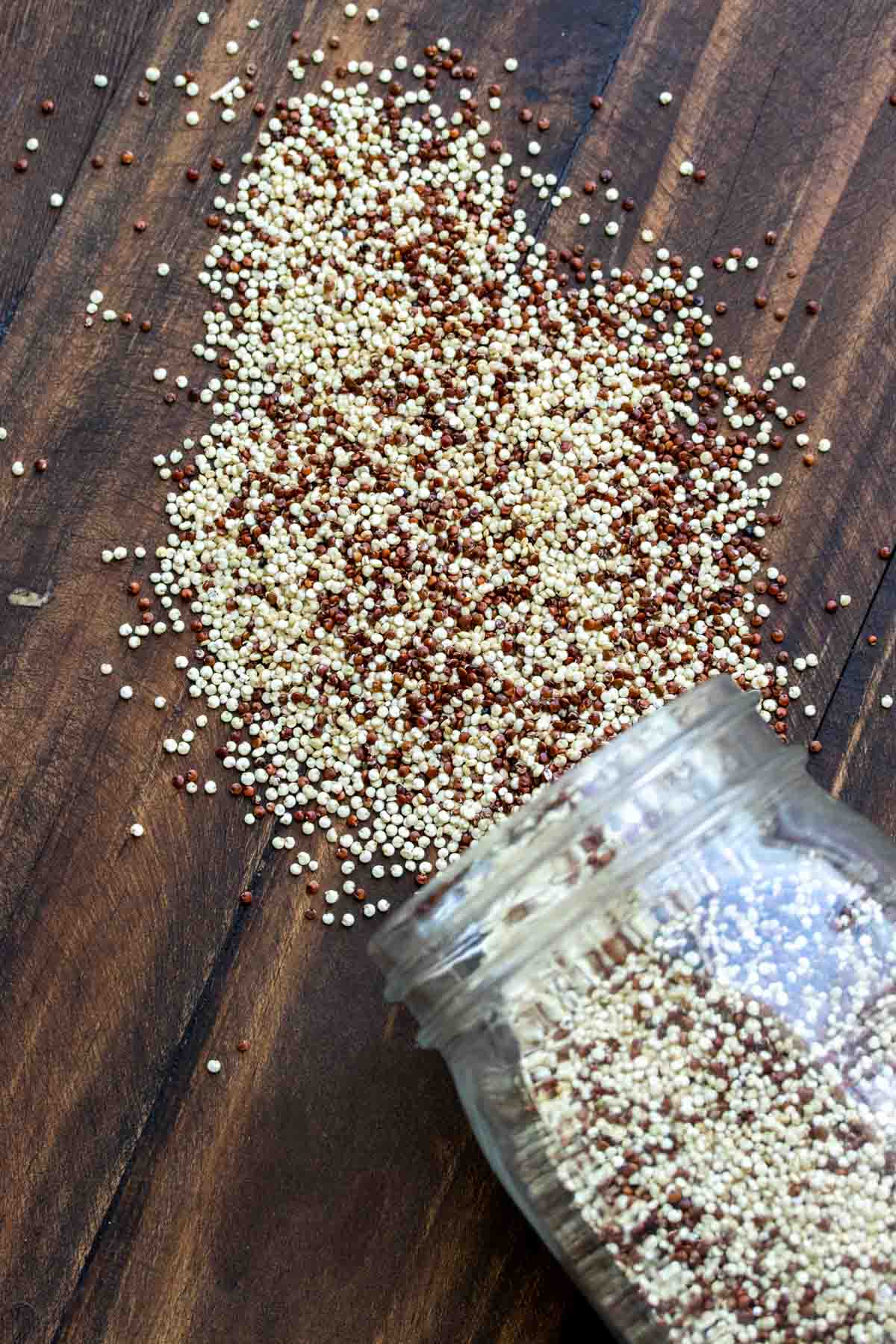 Dry quinoa spilling from a glass jar onto a wooden surface.