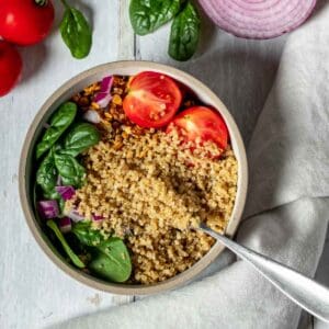 Quinoa with spinach and tomatoes in a white bowl next to more tomatoes and spinach and a red onion on a white wooden surface.