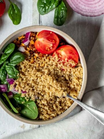 Quinoa with spinach and tomatoes in a white bowl next to more tomatoes and spinach and a red onion on a white wooden surface.