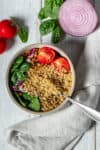 Top view of a white bowl with quinoa, tomatoes and spinach and a spoon in it on a greyish towel.