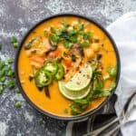 A black bowl on a grey rock background with an orange colored soup with veggies and chickpeas and toppings.