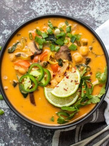 A black bowl on a grey rock background with an orange colored soup with veggies and chickpeas and toppings.