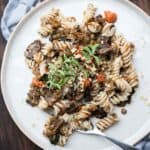 A silver fork on a cream plate with rotini pasta mixed with lentil bolognese and a sprig of thyme on top.