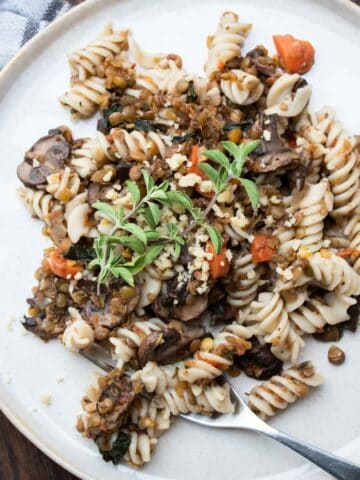 A silver fork on a cream plate with rotini pasta mixed with lentil bolognese and a sprig of thyme on top.