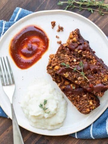 Sliced lentil meatloaf on a white plate with mashed cauliflower and barbecue sauce.