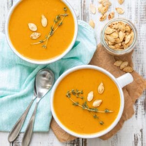 Butternut squash soup in two white soup mugs on a white wooden surface with thyme and pumpkin seeds on the soup.