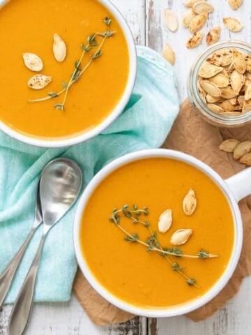 Butternut squash soup in two white soup mugs on a white wooden surface with thyme and pumpkin seeds on the soup.