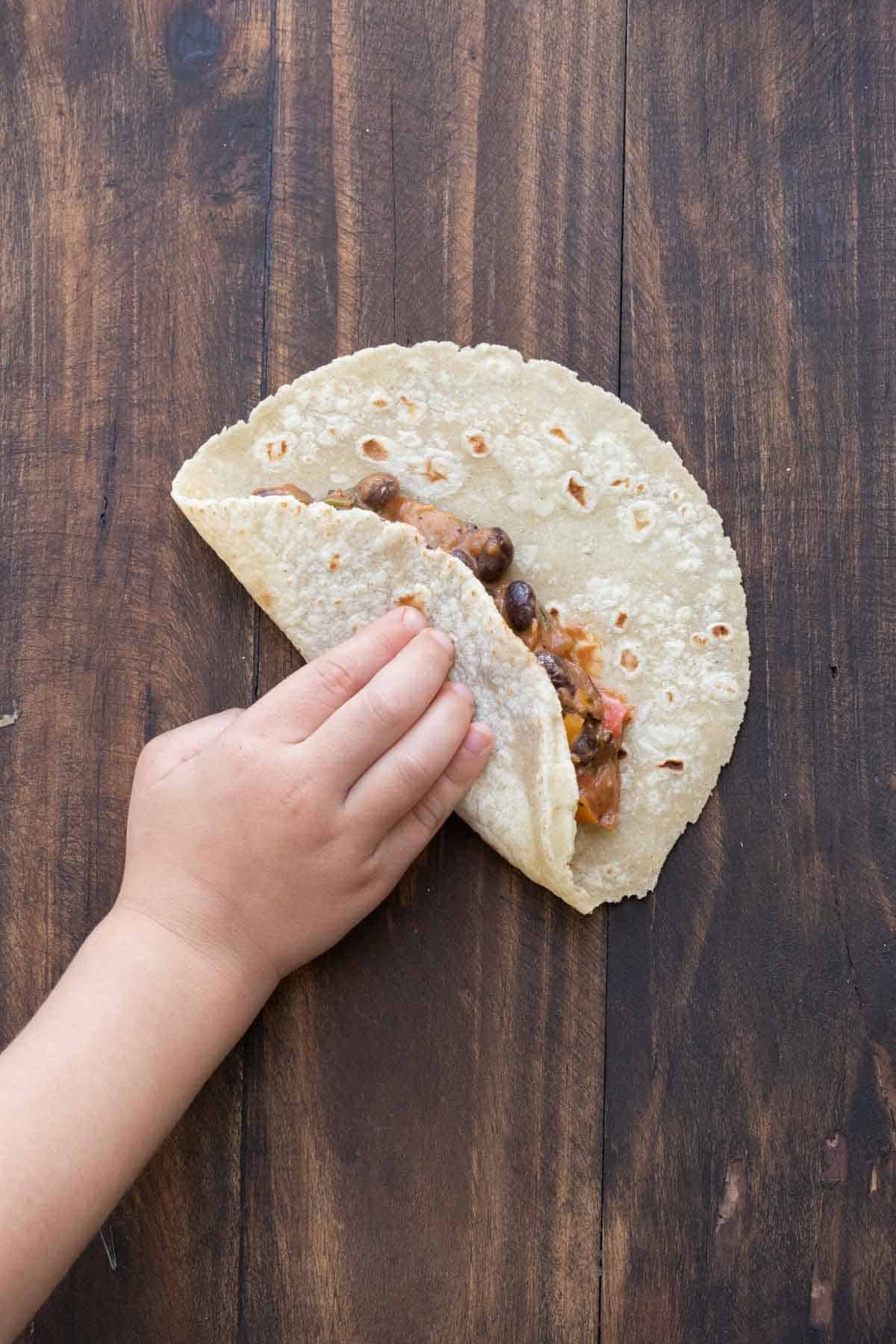 A kids hand folding over a tortilla filled with bean veggie mix.