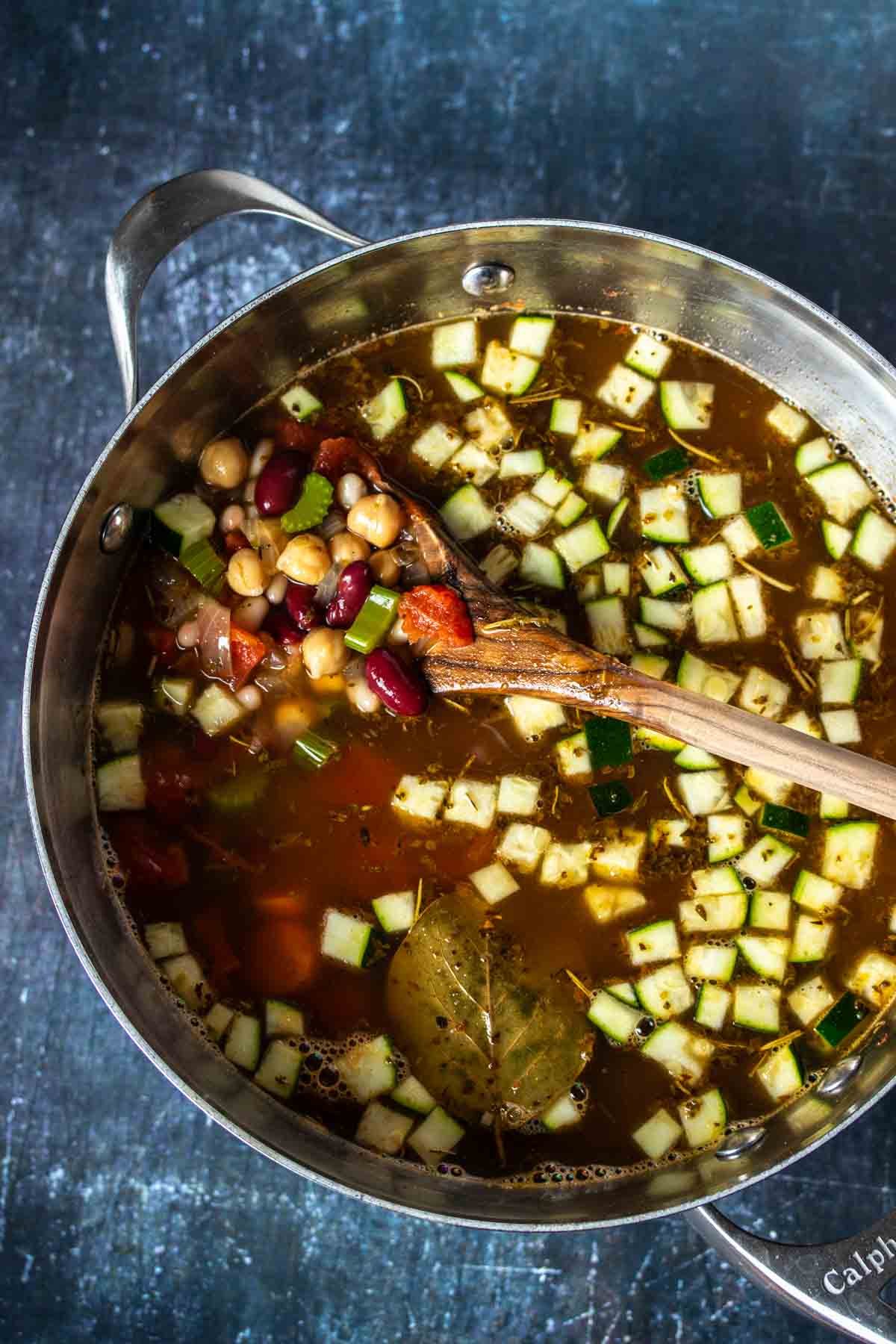 A vegetable and bean broth soup in a metal soup pot being mixed by a wooden spoon.