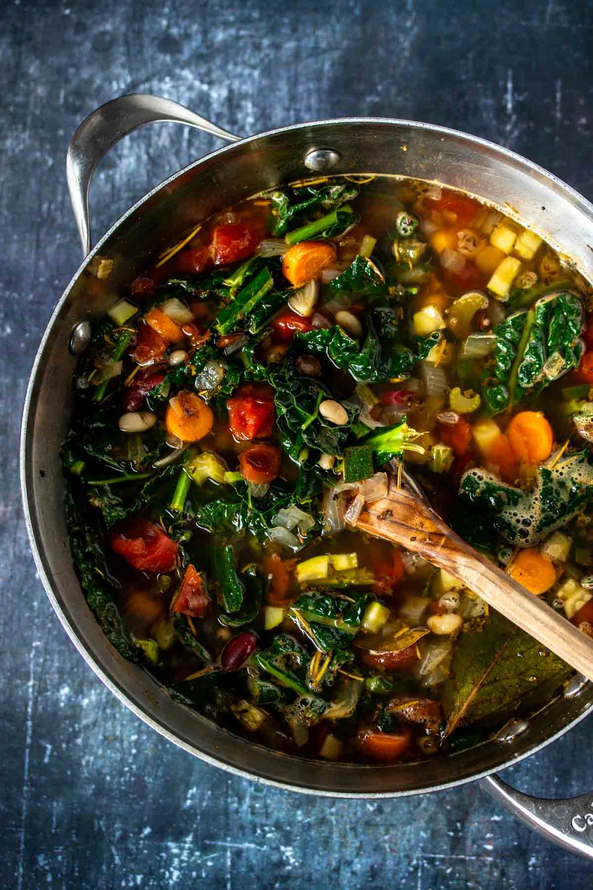 Kale put into a broth based soup with vegetables and beans and a wooden spoon mixing it.