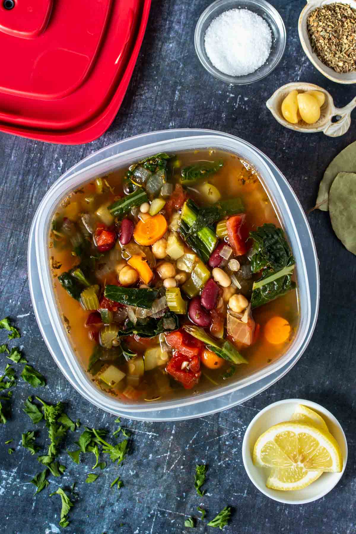 A vegetable bean soup in a plastic container with a red lid sitting next to it.