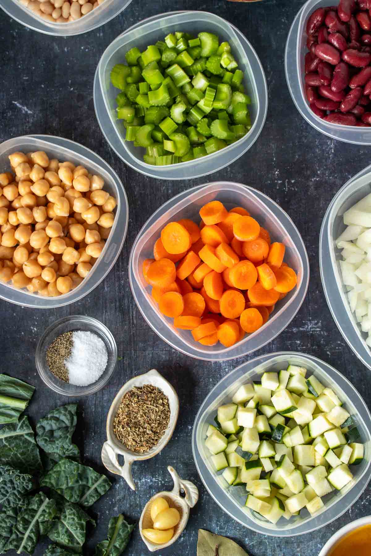 Vegetables and beans in plastic containers next to spices, garlic and kale on a dark surface.