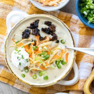 Cream bowl with creamy soup inside topped with sour cream, cheese, scallions and bacon.