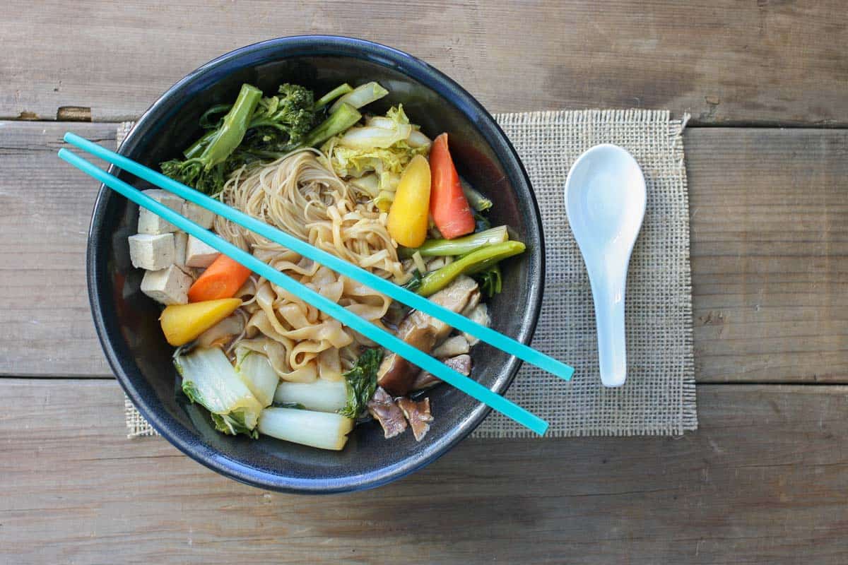 Top view of a black bowl with veggies and noodles in a broth with a white soup spoon next to it.