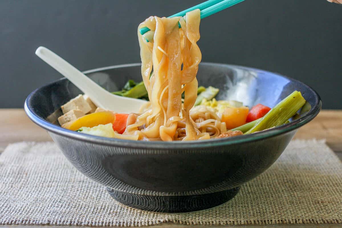 Turquoise chopsticks holding noodles coming out of a bowl of veggie noodle soup.