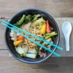 A black bowl with a veggie noodle soup in it on a piece of burlap and a white spoon next to it.