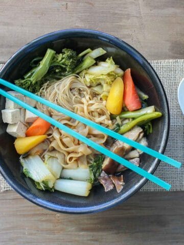 A black bowl with a veggie noodle soup in it on a piece of burlap and a white spoon next to it.