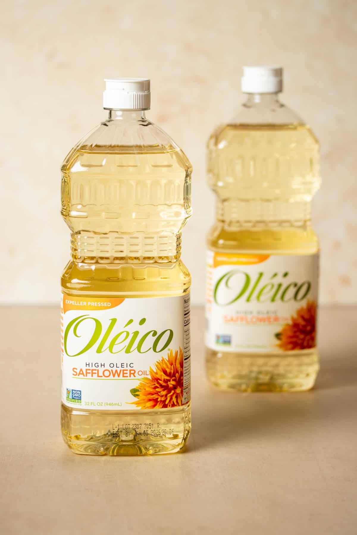 Two bottles of oil with white labels sitting one in front of the other on a tan surface.