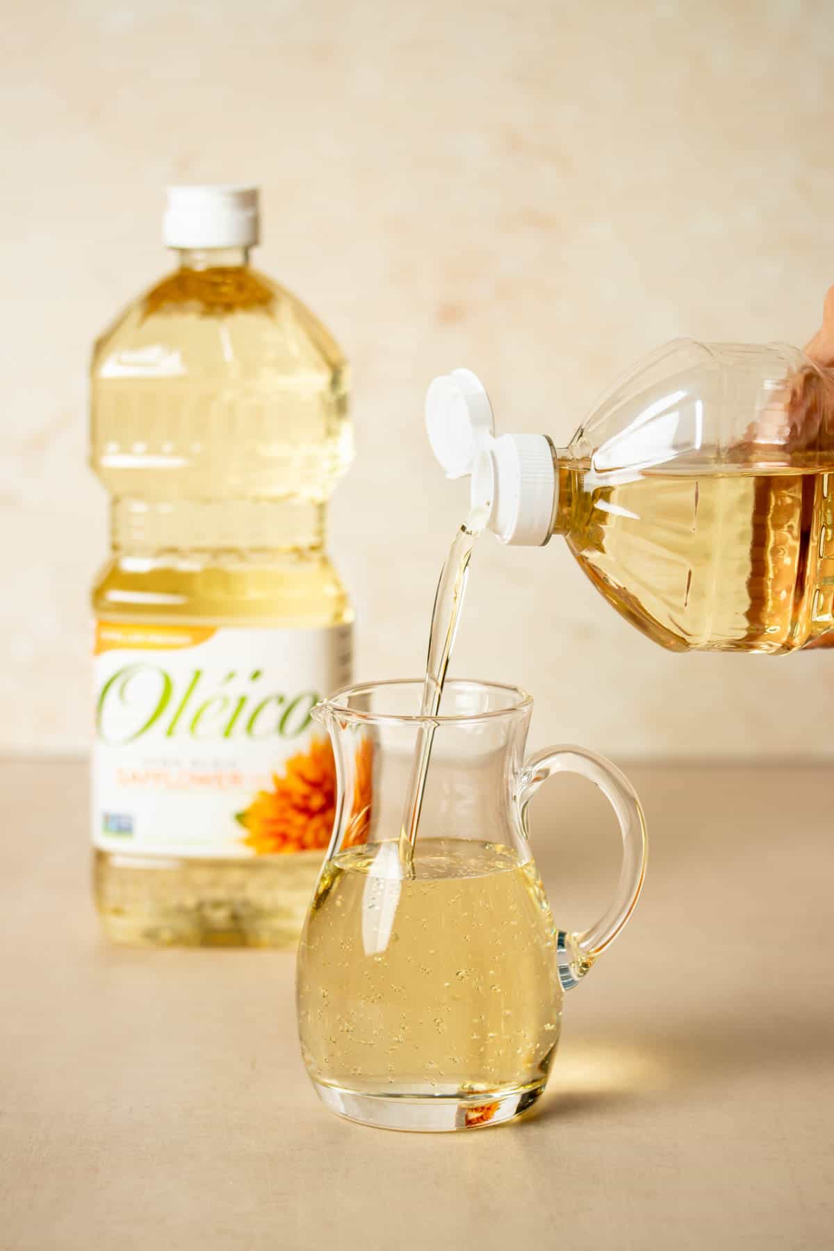 A hand pouring oil from a bottle into a small glass pitcher with another bottle of oil in the background.