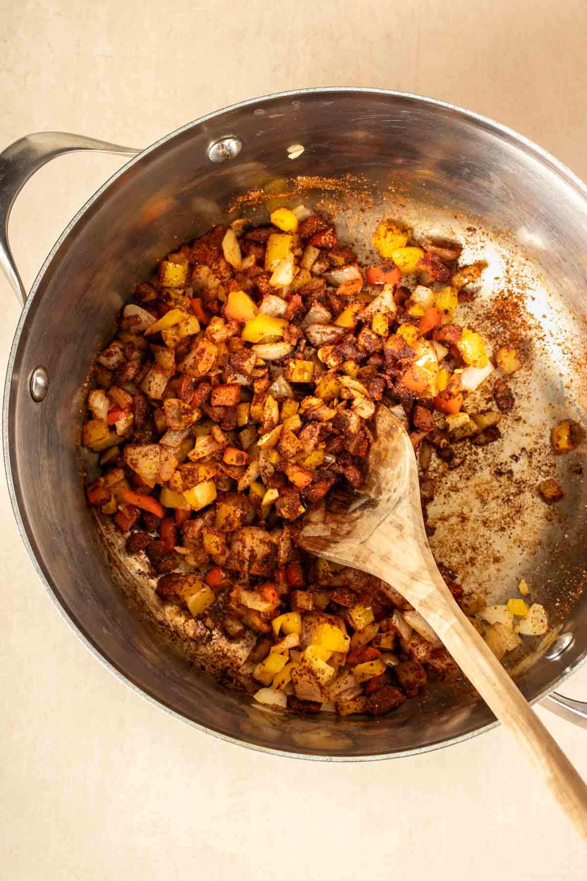 A wooden spoon stirring chopped peppers and onion mixed with red seasonings in a metal pot.