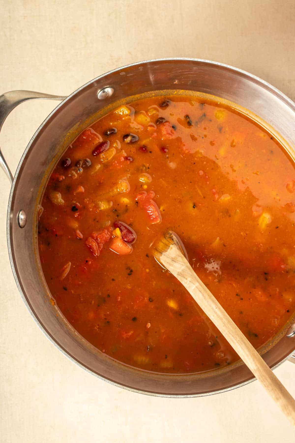 A pot with a red broth and beans and veggies in it being mixed with a wooden spoon.